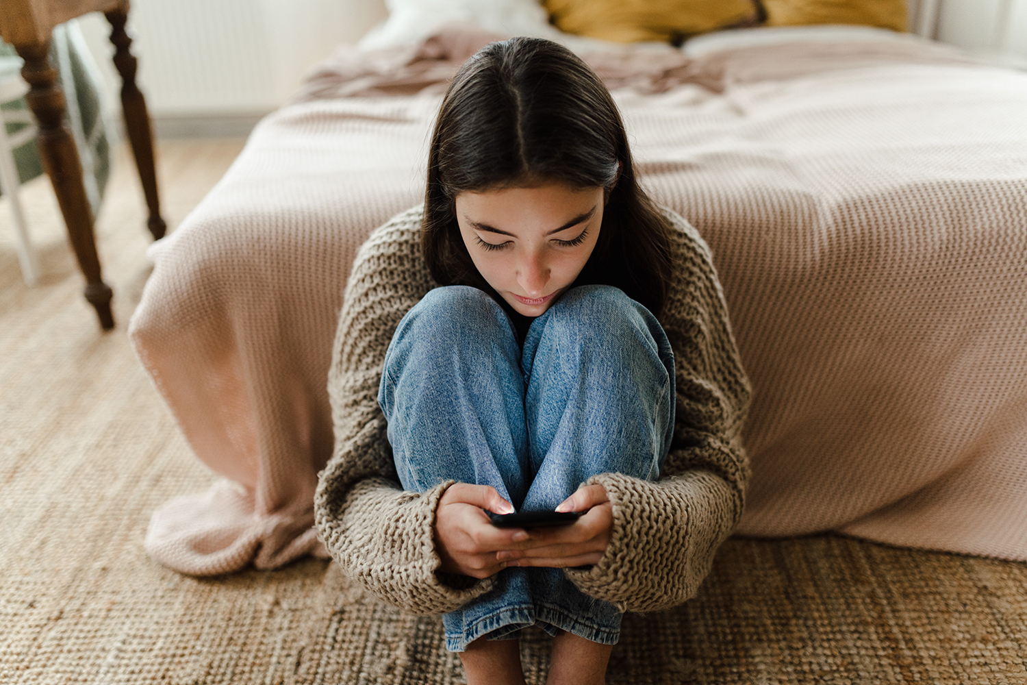 A pensive, teenage girl uses her phone while sitting on the floor in a bedroom.